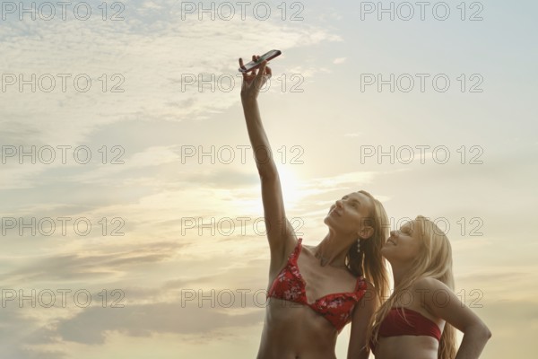 Two friends in swimwear stand on the beach, smiling as they take selfies. The sun sets behind them, casting a warm glow across the sky. Waves gently lap at the shore, creating a relaxing atmosphere