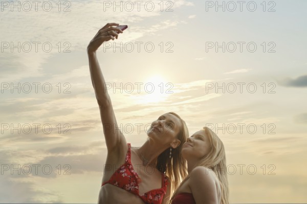 Two friends capture a joyful moment taking a selfie with the sun setting behind them. Dressed in bikinis they take photos to remember their vacation by the water