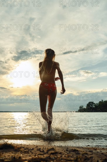 A woman in swimwear joyfully running into a tranquil lake at sunset, with golden rays reflecting on the water. The sandy shore provides a warm backdrop as clouds drift in the sky
