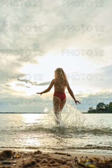 A young woman in a red swimsuit joyfully runs into the lake, creating splashes as she embraces the refreshing water. The setting sun casts a warm glow across the sky and water