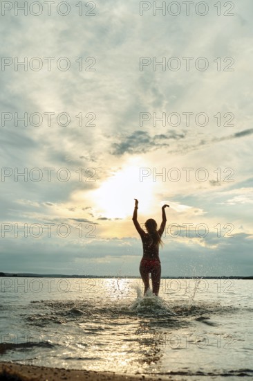 A person stands in the shallow water with arms raised, enjoying the sunset. The sky is filled with soft clouds, while the sun casts a warm glow over the tranquil water