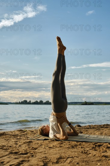 A woman performs shoulder blade stand with her legs raised high at a lakeside on a sandy shore. The sun sets behind her, casting a warm glow over the tranquil water and surrounding landscape
