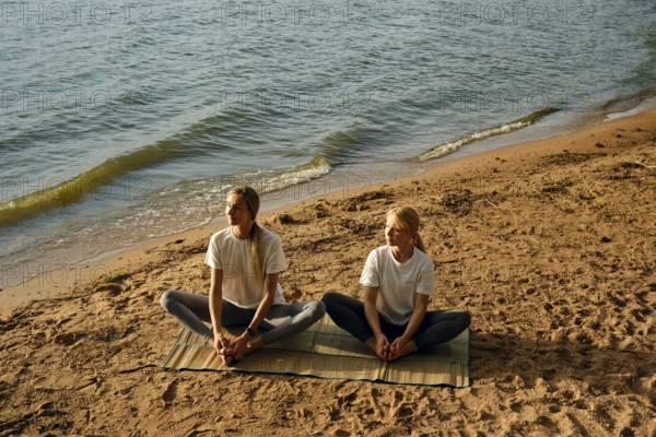 Two women are sitting on a mat by the water's edge and doing yoga poses as the sun begins to set. The sandy beach and gentle waves create a serene atmosphere for their practice