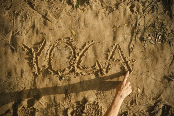 A person draws the word yoga in the sand using her finger. The setting is a sunny beach, showcasing the connection between nature and mindfulness, inviting relaxation and calmness