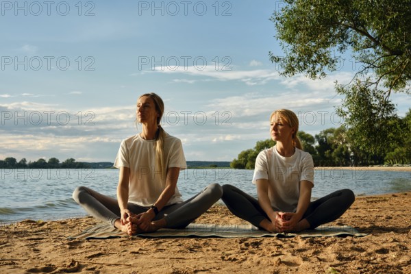 Two women in comfortable clothing are practicing yoga on the beach by a tranquil lake. They sit cross-legged on a mat, enjoying the peaceful natural surroundings and the soft sunlight