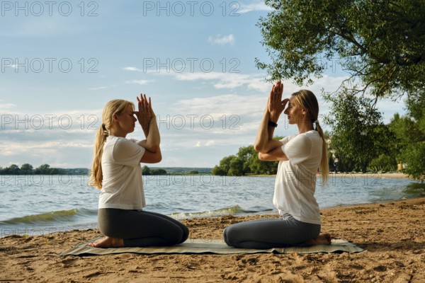 Two women sit on a mat in front of each other by the water, practicing yoga. They perform a meditative pose with hands raised, surrounded by nature as the sun sets behind them