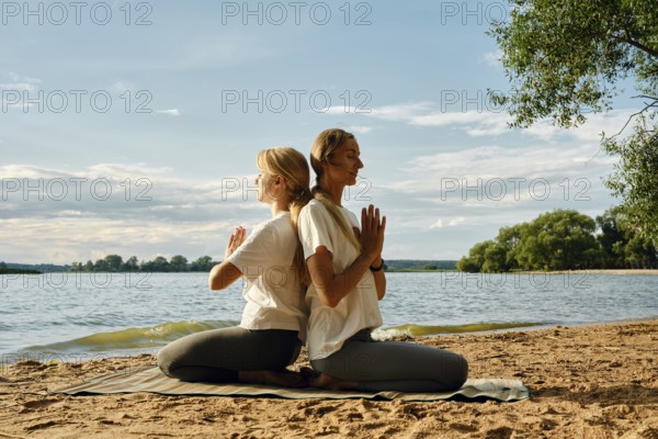 Two women sit back-to-back on a sandy lakeshore, practicing yoga under the warm evening sun. They appear focused and serene, enjoying the peaceful atmosphere created by the water and trees
