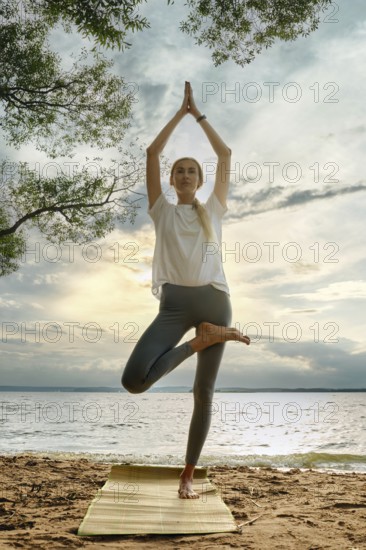 A woman performs a yoga pose on a mat at the water's edge, surrounded by nature. The sky glows with sunset colors, reflecting tranquility and calmness in the atmosphere