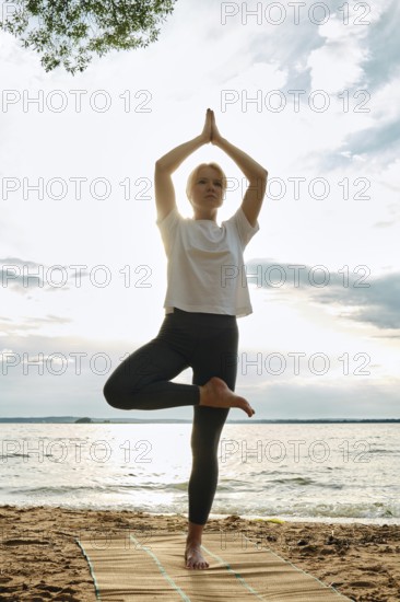 A woman performs yoga on a sandy beach, balancing in a tree pose with arms raised towards the sky. The sun sets behind, casting a warm glow over the peaceful water and clouds