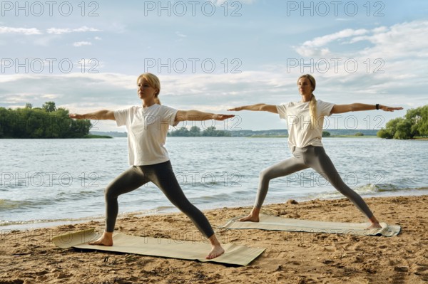 Two women perform yoga warrior pose on mats at a sandy beach by a calm lake. The sun is shining brightly, illuminating their focused expressions as they engage in their practice amidst natural beauty