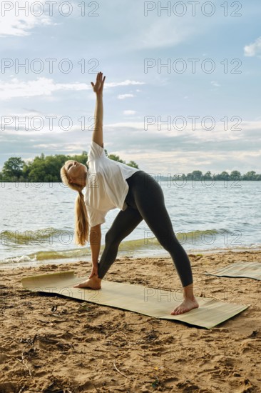 A woman stretches into a side angle pose on a yoga mat placed on the sandy shoreline