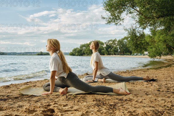 Two women engage in yoga poses on the sandy beach beside a calm lake. The sun shines brightly, illuminating the trees, and creating a serene atmosphere perfect for relaxation and mindfulness