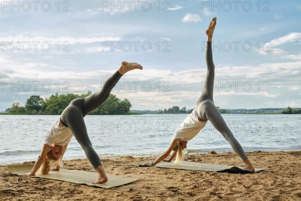 Two women practice yoga on mats along a sandy lakeside during the early evening. They rest their hands on the mat and pull their leg up