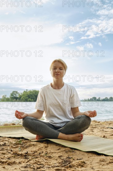 A woman sits cross-legged on a mat by the lake, eyes closed in meditation. The sun shines down on her as gentle waves lap at the shore, surrounded by lush greenery and blue skies