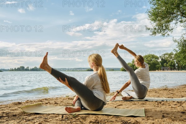 Two women practice yoga on mats by the lake, enjoying the sunny afternoon and calm waters. They showcase various poses while surrounded by trees and beautiful clouds in the sky