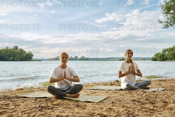 Two women are sitting cross-legged on yoga mats by the riverbank, practicing yoga with serene expressions. The sun shines brightly above, creating a peaceful atmosphere around them