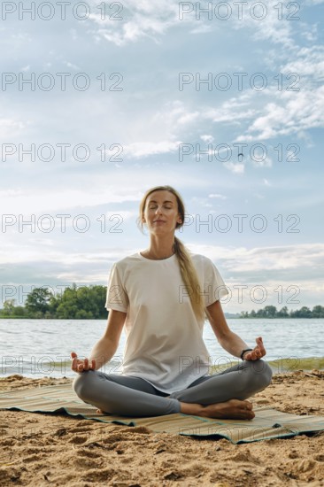 A woman sits cross-legged on a mat by the lake, practicing yoga in a serene environment. The sun shines brightly, and the sky is mostly clear, creating a peaceful atmosphere for mindfulness