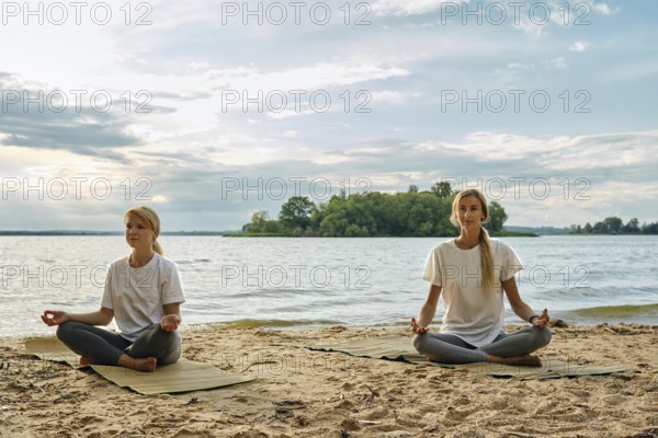 Two women sit cross-legged on yoga mats, meditating by a lake. Soft waves lap against the sandy shore as clouds drift overhead, creating a tranquil atmosphere