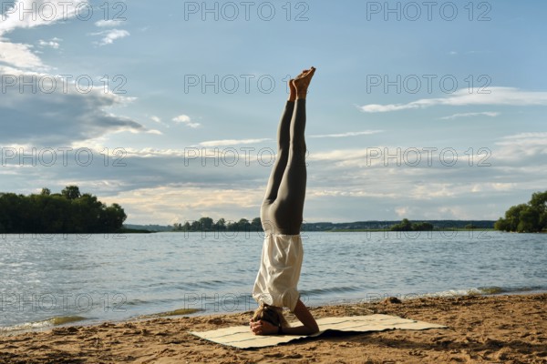 A woman performs a headstand yoga pose on a sandy beach beside a tranquil river. The sun sets in the background, casting warm hues across the sky and reflecting on the water
