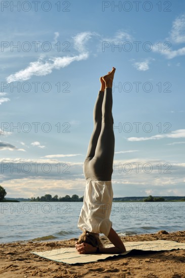 A woman balances in a headstand on a yoga mat on the sandy shoreline of a lake during sunset. The sky is bright blue with scattered clouds, and the surrounding landscape is serene and natural