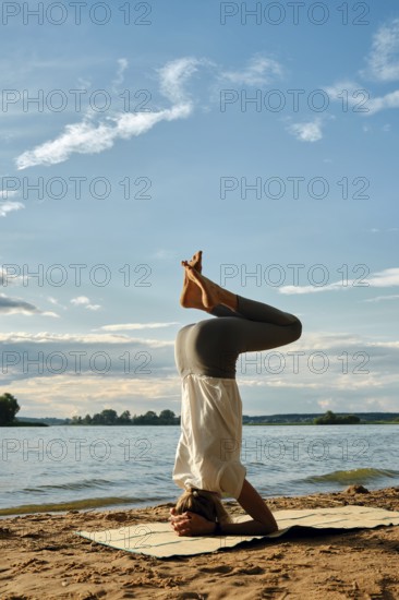 A slender woman performs a headstand yoga pose on a mat in the sand near a tranquil lake. The backdrop features a cloudy sky illuminated by the soft hues of dusk, creating a peaceful atmosphere