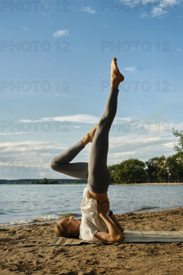 A dedicated individual performs a yoga pose on a sandy beach by the tranquil lake. The sun shines down, casting a warm glow, while lush trees line the water's edge, creating a peaceful atmosphere