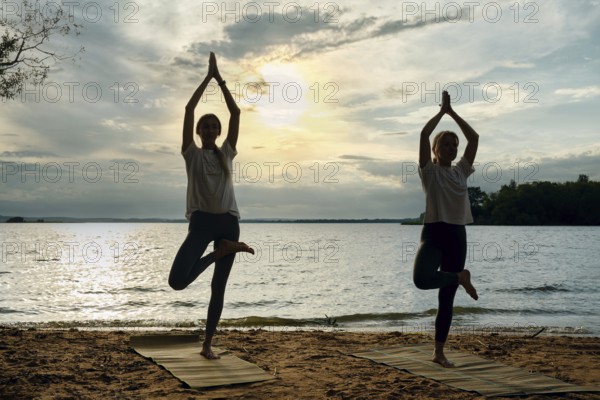 Silhouettes of two women performing yoga poses on mats along a sandy lakeshore as the sun sets, casting a warm glow over the water and creating a peaceful atmosphere