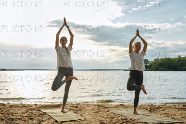 Two women perform yoga poses on mats at the beach during sunset. They are surrounded by a tranquil lake and a beautiful sky, promoting relaxation and mindfulness in nature