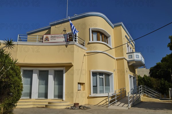 A modern yellow building with balconies and Greek flag, artistically designed, Armando Bernabiti, Historicism, Rationalism, Art Deco, Rationalism, Rodolfo Petraco, Fascism, Portolago, Lakki, Leros, Dodecanese, Greek Islands, Greece