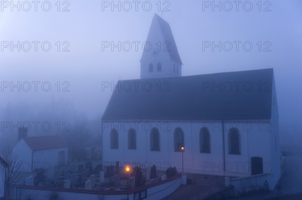St. Martin's parish church with cemetery, orange-coloured street lamps in the morning fog in front of sunrise, first light, blue hour, Marzling, Freising, Upper Bavaria, Bavaria, Germany