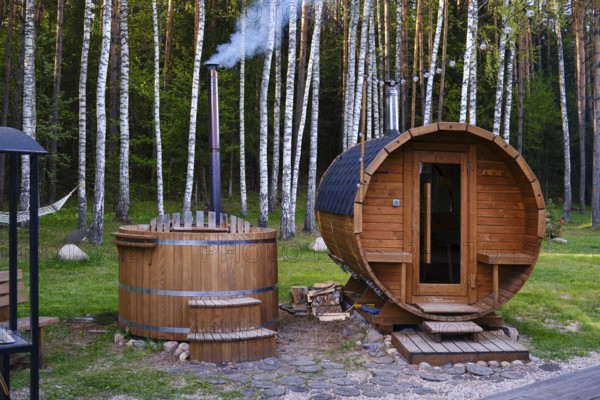 A wooden sauna and a hot tub are nestled in a lush forest. The hot tube emits smoke from its chimney, and a hammock nearby invites relaxation amid nature