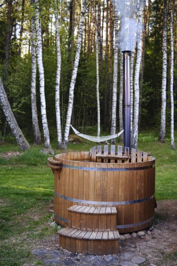 A rustic wooden hot tub sits in a peaceful clearing, surrounded by tall trees and green grass. A gentle plume of steam rises from the chimney, suggesting a warm soak