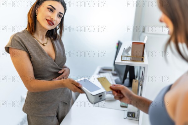 Beautician holding a pos terminal accepting contactless payment from a female customer with a credit card inside a beauty salon