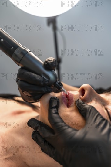 Beautician wearing black gloves applying permanent makeup on lips using a tattoo machine in a beauty salon