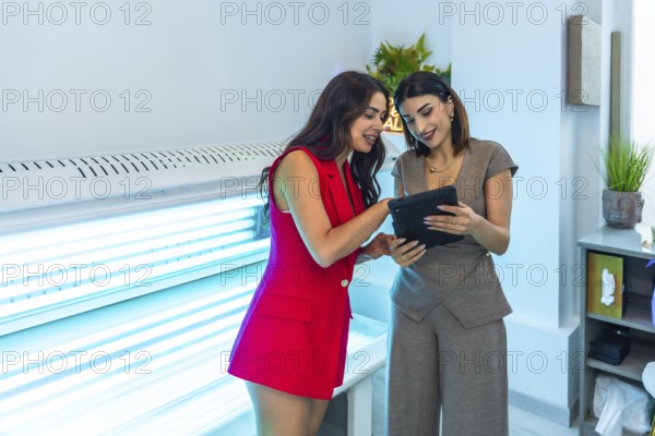 Two beauticians are consulting a digital tablet, discussing treatment options near a solarium, inside a beauty salon or aesthetic center