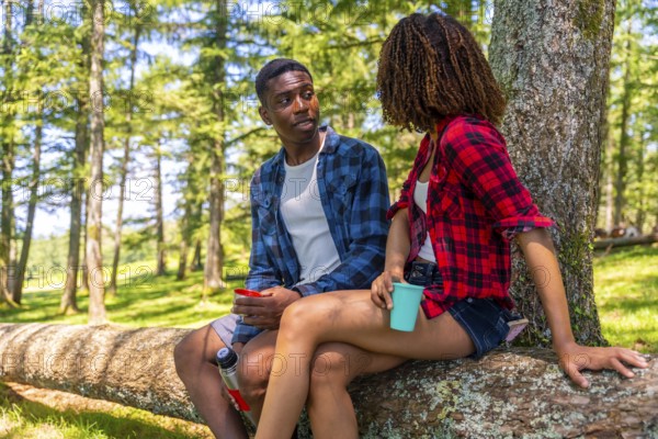 Two hikers are taking a break in the woods, sitting on a fallen tree trunk, enjoying a conversation and drinks from reusable cups