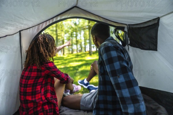 Two tourists sitting inside their tent, excitedly pointing at something intriguing in the lush forest while enjoying a camping adventure