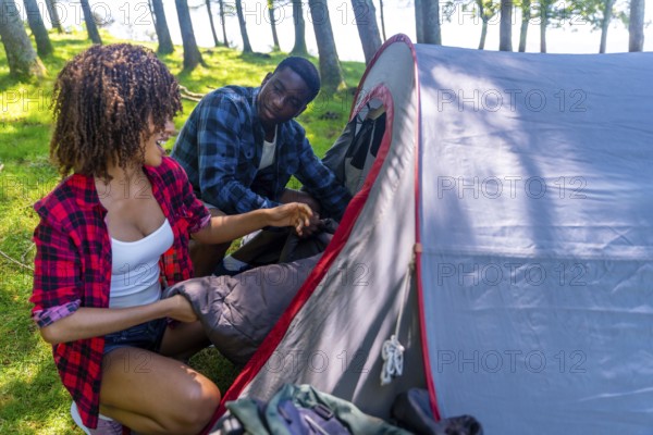 Young black couple enjoying the outdoors while setting up a camping tent in the forest, placing a sleeping bag inside with smiles