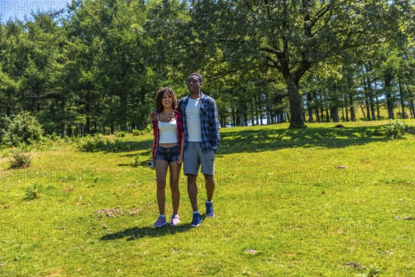 Young multi ethnic couple enjoying a sunny summer day, walking hand in hand through a lush green meadow surrounded by trees and nature