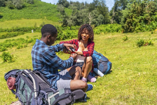 Young couple of hikers taking a break on a grassy hillside, sharing refreshments from a thermos during a sunny day hike