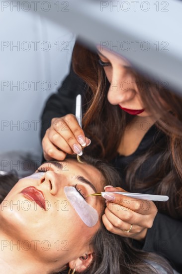 Beautician carefully applying individual false eyelashes on client's upper eyelid using tweezers in a professional beauty salon