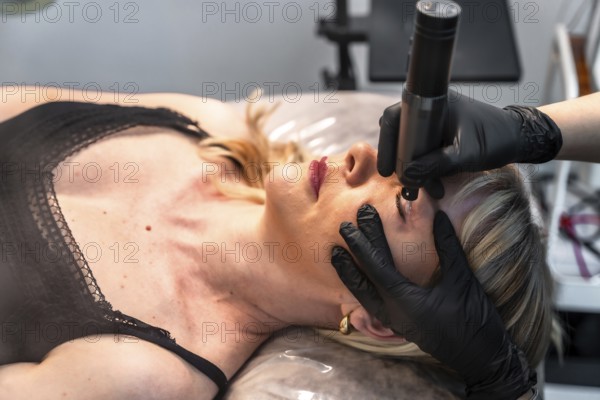 Beautician applying permanent eyeliner makeup on upper eyelid of client lying on bed in beauty salon