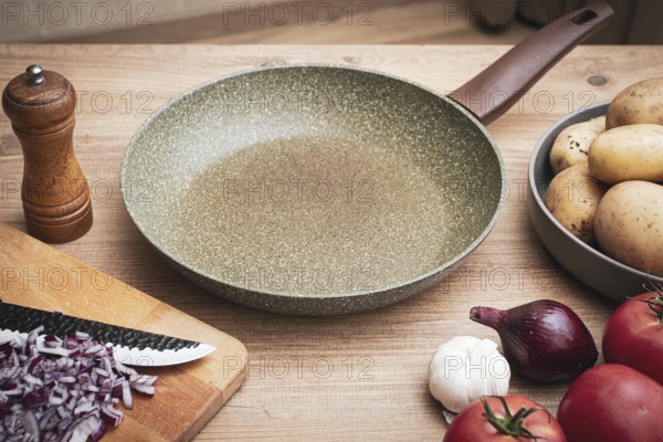Fresh ingredients including potatoes, garlic, tomatoes, and onions are arranged on a wooden table alongside a frying pan and a knife for cooking preparation