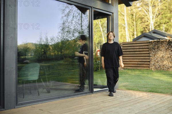 Two boys are walking out of a sleek, contemporary house showcasing large glass doors. The surrounding area features lush greenery and organized wood stacks, creating a peaceful atmosphere