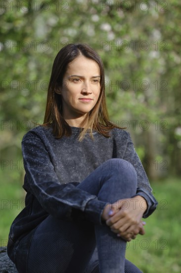 A woman sits quietly in an outdoor area, surrounded by lush greenery and soft light. She appears contemplative while sitting cross-legged on a rock, radiating a serene presence amidst nature