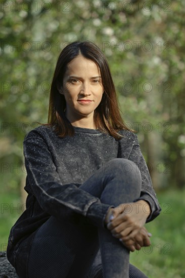 A young woman with long dark hair sits comfortably on a rock, wearing a casual sweater and leggings. She looks directly at the camera, with soft sunlight filtering through trees in the background