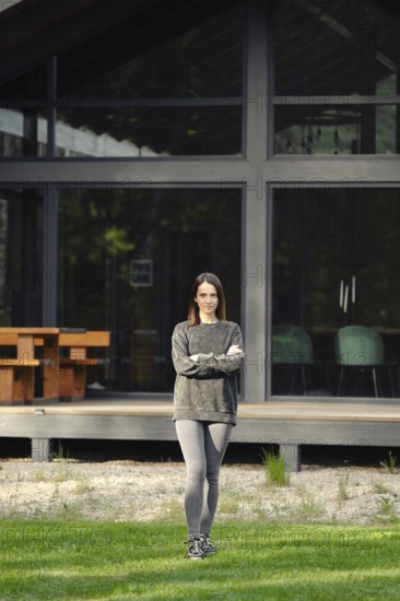 A young woman stands crossing her hands on the lawn in front of her modern house with large glass windows