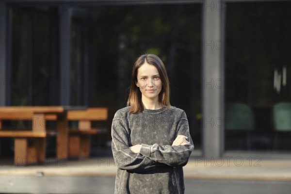 A young woman with shoulder-length hair stands confidently with her arms crossed outside a sleek, modern building