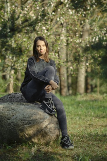 A young woman with long hair sits thoughtfully on a large rock in a peaceful forest. She wears casual clothing, enjoying the tranquility of nature. Sunlight filters through the trees