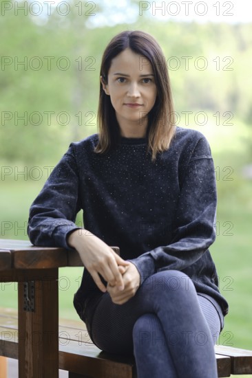A woman is seated on a wooden bench, dressed in a black sweatshirt and leggings. She gazes directly at the viewer with a calm expression, surrounded by lush greenery in the background
